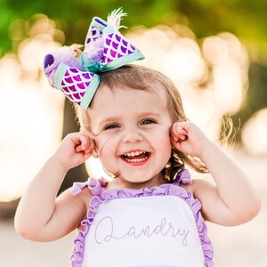 May include: A young girl wearing a light blue and white dress with purple ruffles and the name "Landry" embroidered on the front. She is wearing a purple and teal bow with a mermaid scale pattern in her hair. She is smiling and has her hands over her ears.