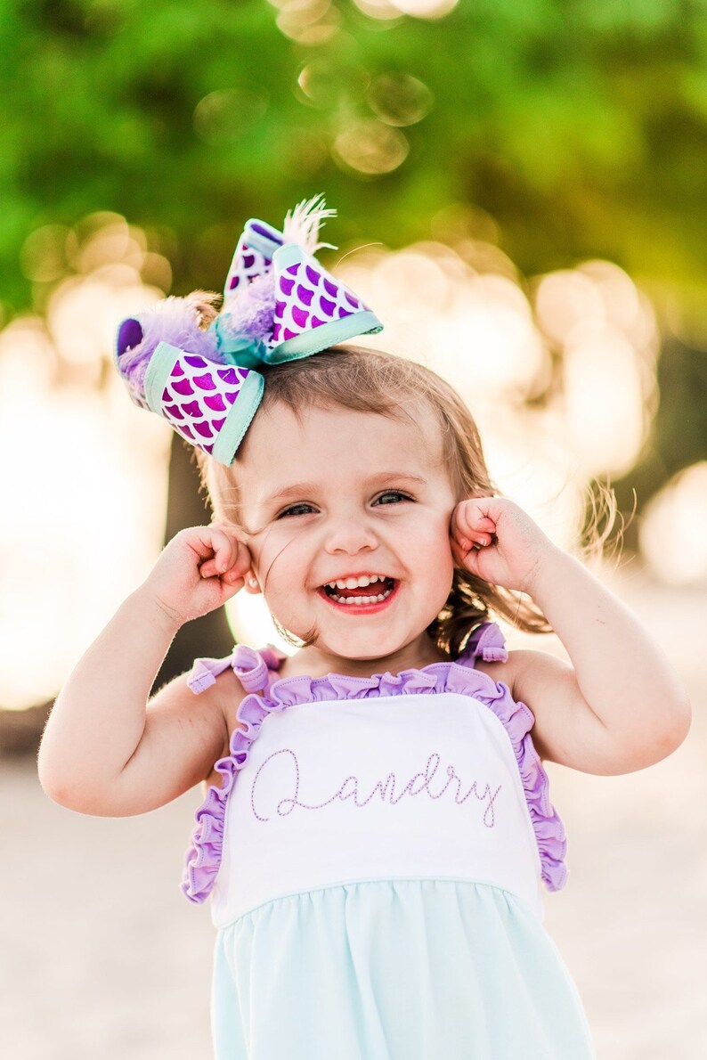May include: A young girl wearing a light blue and white dress with purple ruffles and the name "Landry" embroidered on the front. She is wearing a purple and teal bow with a mermaid scale pattern in her hair. She is smiling and has her hands over her ears.