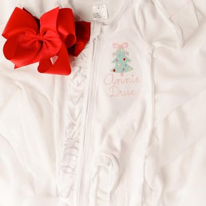 Puede incluir: Mono de beb&eacute; blanco con un dise&ntilde;o de &aacute;rbol de Navidad y el nombre "Annie Drue" bordado en hilo rosa. Un gran lazo rojo se coloca junto al mono. El mono tiene cierre de cremallera y ribete de volantes.