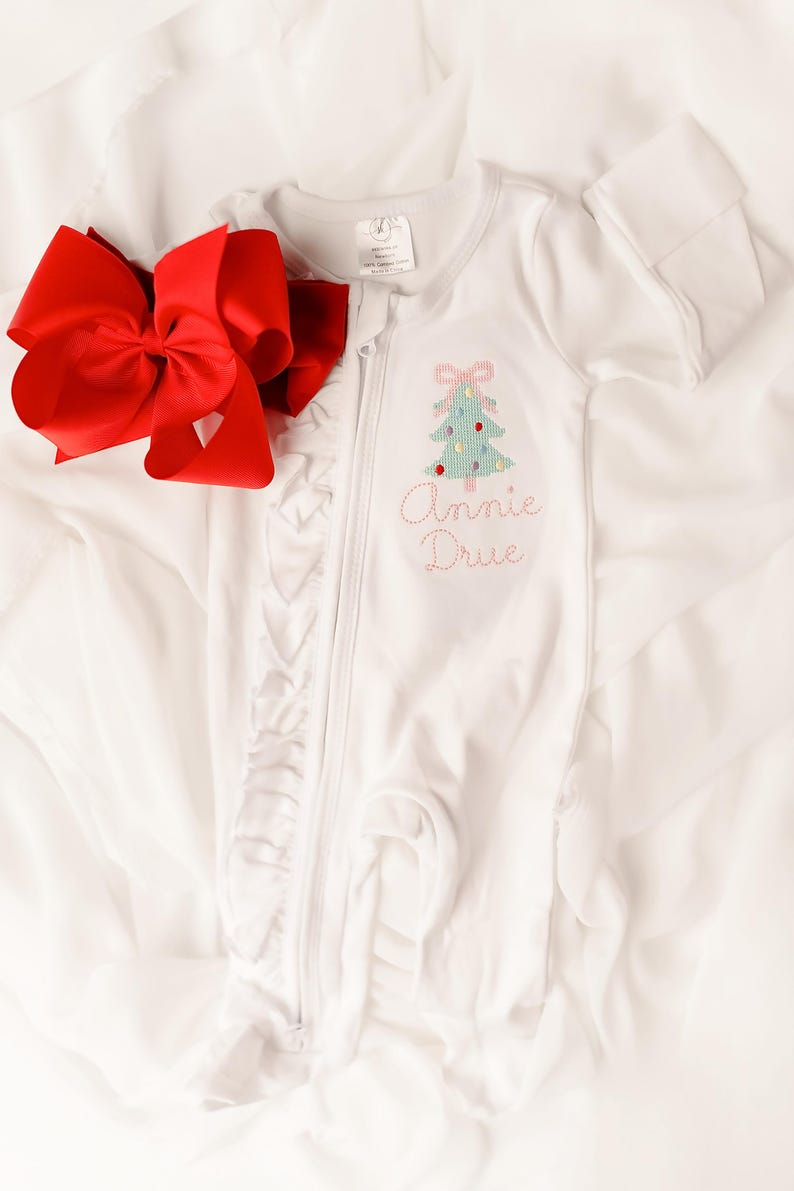 Puede incluir: Mono de beb&eacute; blanco con un dise&ntilde;o de &aacute;rbol de Navidad y el nombre "Annie Drue" bordado en hilo rosa. Un gran lazo rojo se coloca junto al mono. El mono tiene cierre de cremallera y ribete de volantes.
