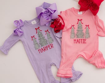 May include: Two long-sleeved rompers in lavender and pink, each featuring an embroidered design of three silver Christmas trees topped with red bows. The lavender romper is personalized with the name HARPER, and the pink romper with HATTIE.