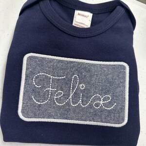 May include: A navy blue bodysuit with a white embroidered rectangle patch that says "Felice" in a cursive font.