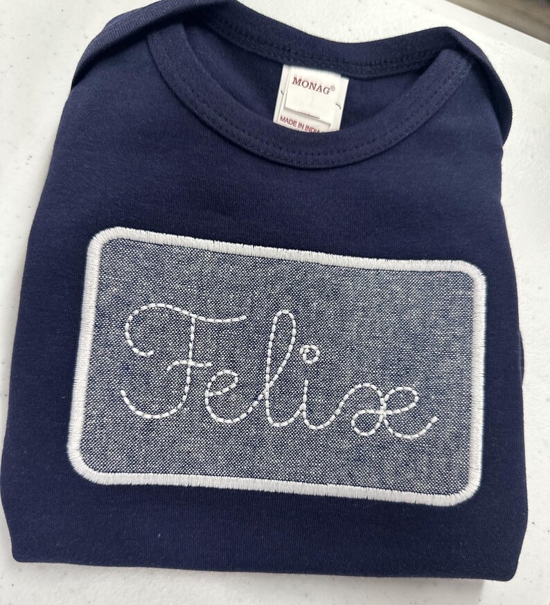 May include: A navy blue bodysuit with a white embroidered rectangle patch that says "Felice" in a cursive font.