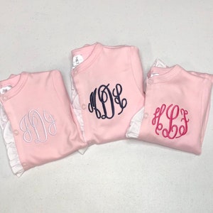 May include: Three pink baby onesies with white ruffles around the neckline. Each onesie has a different monogram embroidered on the front. The monograms are in white, navy blue, and pink.