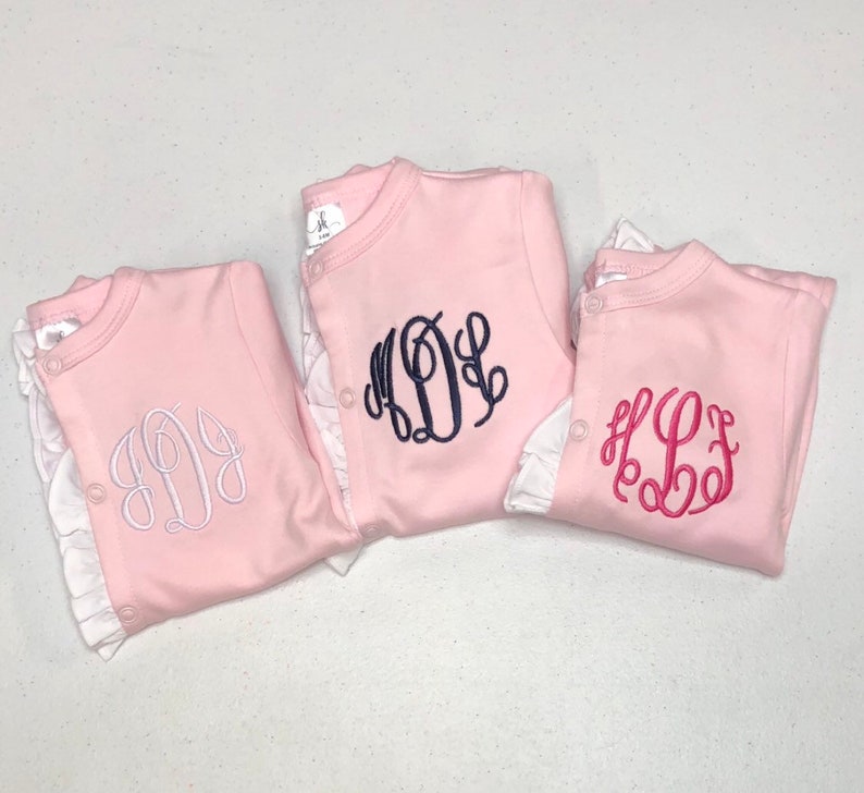 May include: Three pink baby onesies with white ruffles around the neckline. Each onesie has a different monogram embroidered on the front. The monograms are in white, navy blue, and pink.