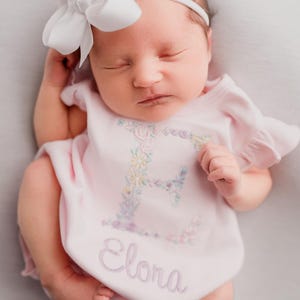 May include: A newborn baby wearing a pink onesie with the name "Elora" embroidered on it. The onesie has a floral "E" design. The baby is also wearing a white headband with a large white bow.