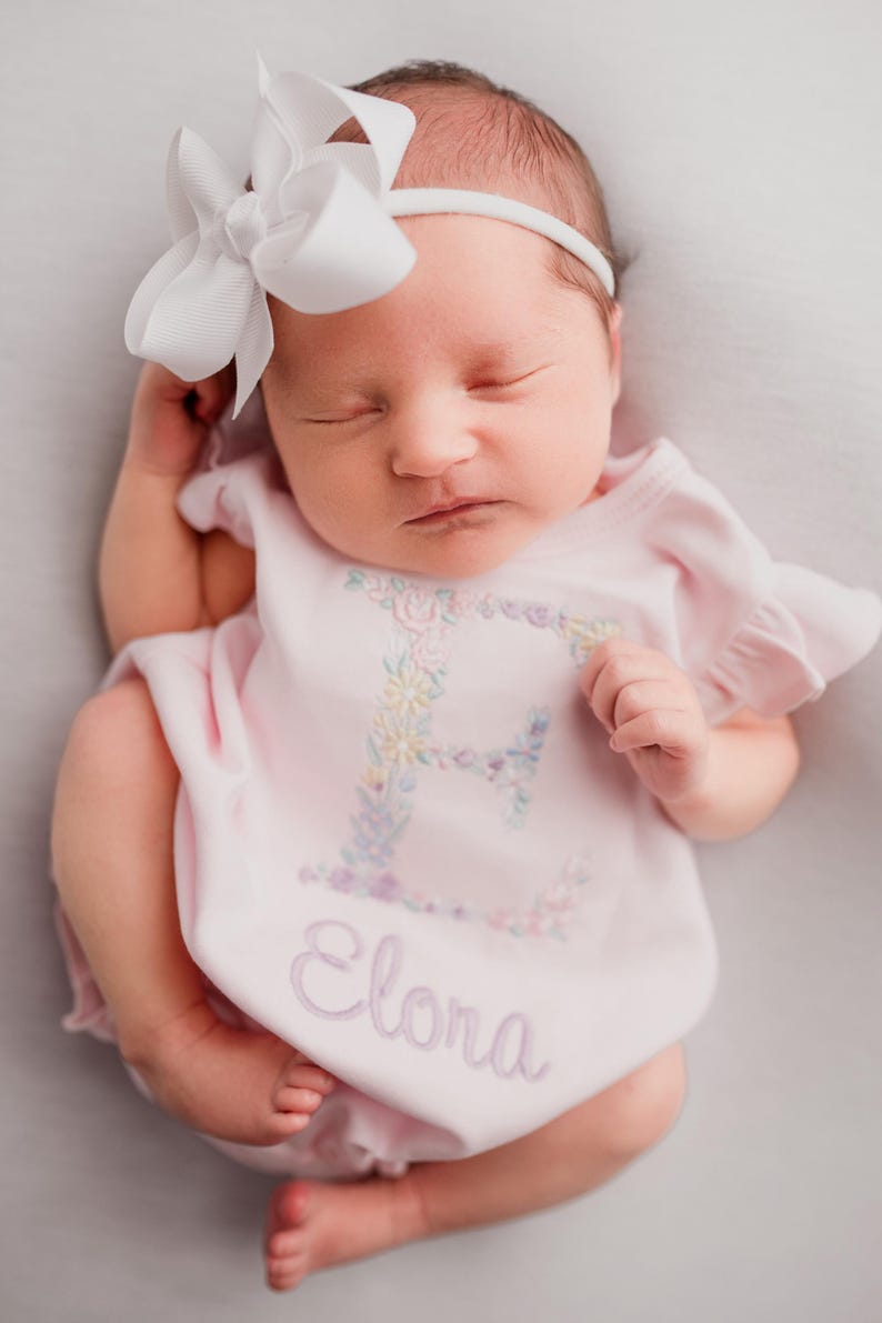 May include: A newborn baby wearing a pink onesie with the name "Elora" embroidered on it. The onesie has a floral "E" design. The baby is also wearing a white headband with a large white bow.