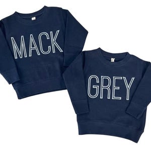 Puede incluir: Dos sudaderas azul marino. Una tiene el nombre "MACK" en letras blancas. La otra muestra "GREY" en letras blancas. Ambas tienen cuello redondo y mangas largas, ideales para ni&ntilde;os.