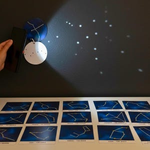 Puede incluir: Un proyector de constelaciones, sostenido por una persona, proyecta estrellas sobre una pared oscura. El proyector tiene un diseño azul y blanco. Debajo hay tarjetas de constelaciones con dibujos de líneas amarillas sobre un fondo azul.