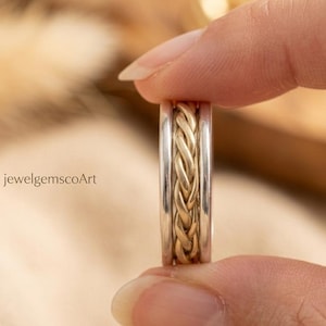 May include: A close-up of a two-tone ring with a braided gold center and silver bands. The ring is held between two fingers. The text "jewelgemscoArt" is visible in the background.