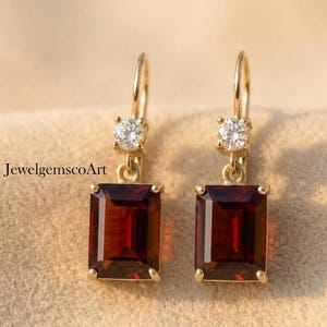 May include: Gold-coloured drop earrings with a rectangular, deep red gemstone and a small, round, clear stone in each. The earrings hang from a gold hook. The background is a soft, neutral colour. The text "JewelgemscoArt" is visible in the image.