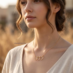 Può includere: Una collana d'oro con tre ciondoli a forma di cuore interconnessi. La collana è indossata da una persona con un top color crema, con uno sfondo sfocato di erba marrone dorato. Il gioiello è delicato ed elegante.