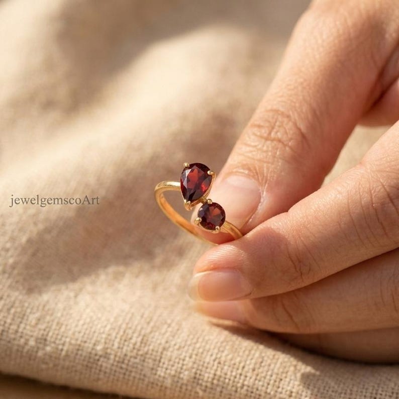 Peut inclure: Une bague en or orn&eacute;e de deux pierres pr&eacute;cieuses rouge fonc&eacute;, l'une en forme de poire et l'autre ronde, dispos&eacute;es en style bypass. L'anneau est fin et en or poli. Le texte jewelgemscoArt est visible &agrave; gauche de l'image, pr&egrave;s de la bague.