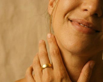 Könnte beinhalten: Ein goldfarbener Ring mit einem glatten, abgerundeten Design. Der Ring wird an einem Finger getragen. Der Ring scheint aus Metall zu sein und hat ein mattes Finish. Das Bild ist eine Nahaufnahme.