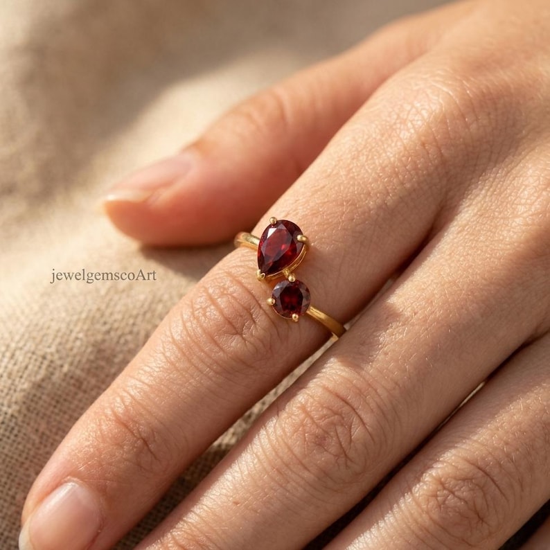 Peut inclure: Une bague en or orn&eacute;e de deux pierres pr&eacute;cieuses grenat rouge fonc&eacute;, l'une en forme de poire et l'autre ronde, mont&eacute;es sur un anneau fin en or. Le texte jewelgemscoArt est visible sur le c&ocirc;t&eacute; gauche de l'image, pr&egrave;s de la bague.