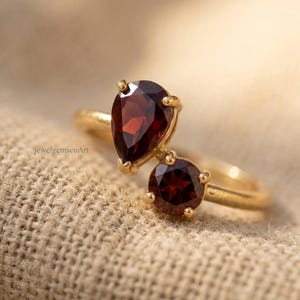 Peut inclure: Une bague en or orn&eacute;e de deux pierres pr&eacute;cieuses grenat rouge fonc&eacute;, l'une en forme de poire et l'autre en taille ronde, mont&eacute;es sur un anneau de style bypass. Le texte jewelgemscoArt est visible sur le c&ocirc;t&eacute; gauche de la bague pos&eacute;e sur un tissu.