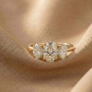 May include: A gold ring with three oval-cut diamonds. The center diamond is larger than the two side stones. The ring is set on a gold band and rests on a tan fabric.