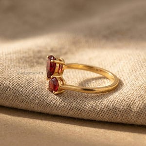 Peut inclure: Une bague en or orn&eacute;e de deux pierres pr&eacute;cieuses rouge fonc&eacute; de taille ovale, dispos&eacute;es en style bypass. L'anneau est lisse et poli, avec le texte jewelgemscoArt visible pr&egrave;s du sertissage. Le design pr&eacute;sente deux pierres rouges.