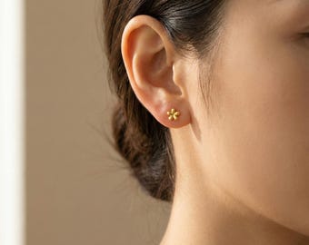 Könnte beinhalten: Nahaufnahme eines Ohrs mit einem kleinen, goldfarbenen Blütenohrstecker. Der Ohrring hat ein zartes Design mit fünf Blütenblättern. Der Hintergrund ist in einem weichen, neutralen Farbton gehalten, der die Details des Ohrrings hervorhebt.