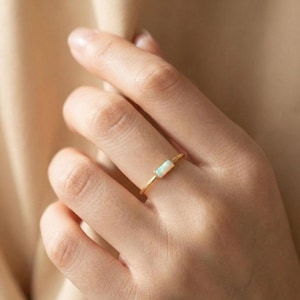 May include: A gold ring featuring a rectangular opal stone. The ring is simple and elegant, with a thin gold band. The opal has a white and iridescent appearance. The ring is shown on a person's finger.
