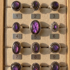Puede incluir: Exhibición de doce anillos de plata con piedras de labradorita morada ovaladas y redondas. Los anillos están dispuestos en una vitrina de madera con etiquetas numeradas. Las piedras tienen una cualidad iridiscente.