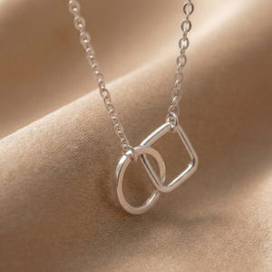 Puede incluir: Collar plateado con una cadena fina y un colgante con un diseño entrelazado de círculo y cuadrado. El colgante es el punto focal, con la cadena que se extiende hacia arriba. El fondo es de color beige neutro.