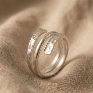Peut inclure: Une bague argentée à la texture martelée. La bague présente un design en spirale, avec deux bandes qui s'enroulent autour du doigt. La bague est posée sur un fond en tissu de ton neutre.