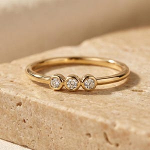 May include: A gold ring featuring three small, clear gemstones. The ring is set on a light-coloured, textured surface. The image is lit to create soft shadows.