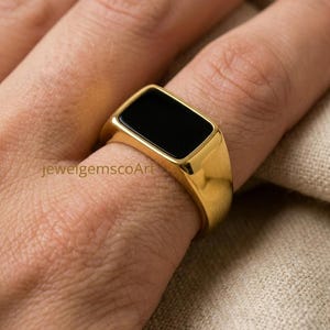 May include: A gold signet ring featuring a rectangular black inlay. The ring has a polished metallic finish and a smooth, flat top surface. The text jewelgemscoArt is visible on the side of the ring band, which is worn on a finger.