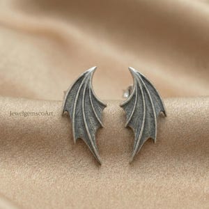 May include: A pair of silver-toned stud earrings shaped like dragon wings. The earrings have detailed veining and a textured surface. The jewelry is displayed on a neutral, satin-like background. The text "JewelgemscoArt" is visible.