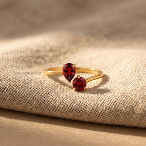 Peut inclure: Une bague ouverte en or orn&eacute;e de deux pierres pr&eacute;cieuses rouge fonc&eacute;, l'une en forme de poire et l'autre ronde, mont&eacute;es sur un fin anneau en or poli. Le texte jewelgemscoArt est visible sur le c&ocirc;t&eacute; gauche de la bague, pr&eacute;sent&eacute;e clairement.