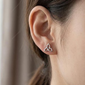 May include: A close-up of a silver-colored stud earring in the shape of a Celtic knot, also known as a triquetra, on an ear. The earring is small and has a simple, elegant design. The background is blurred.
