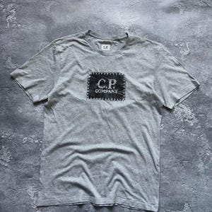Puede incluir: Una camiseta de manga corta de color gris claro con un parche rectangular oscuro en el pecho con el texto C.P. COMPANY impreso en blanco. El parche está unido con costuras en zigzag visibles. Una pequeña etiqueta C.P. COMPANY está en el cuello.