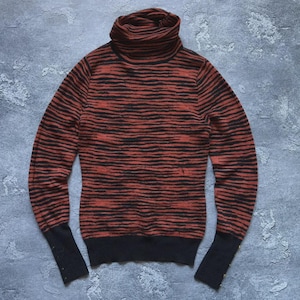 Puede incluir: Un jersey de cuello alto con un patrón de rayas horizontales en tonos rojos y negros. Las mangas y el bajo son de color negro liso. El jersey tiene una silueta ajustada y una textura acanalada.