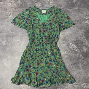 May include: A green short-sleeved dress featuring a vibrant floral pattern in shades of purple, blue, and white. The garment has a V-neckline, a cinched elastic waist, and a ruffled hemline. A small white brand label is visible at the inner neckline.