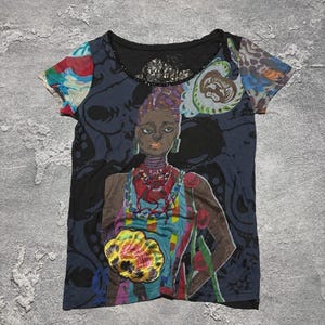 May include: A short-sleeve t-shirt featuring a dark gray base with a large, colorful graphic print of a person wearing a patterned top and jewelry. The design includes abstract shapes, floral elements, and a prominent yellow and orange flower motif.