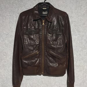 May include: A brown leather jacket with a zip-up front, two chest pockets, and two side pockets. The jacket has a bomber-style bottom and cuffs. The D&G label is visible on the collar.