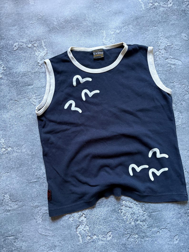 Puede incluir: Camiseta sin mangas azul marino con ribete blanco y un gr&aacute;fico de p&aacute;jaros blancos. La camiseta tiene la etiqueta de la marca "Evisu". La camiseta est&aacute; hecha de un material suave.