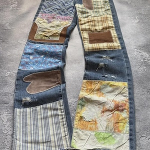 Op de afbeelding: Blauwe denim jeans met patchwork details. Patches bevatten verschillende stoffen met bloemen-, ruit- en abstracte patronen, samen met bruine suède-achtige accenten. De jeans heeft een distressed look met rafelige randen en een klassiek vijf-zakken ontwerp.