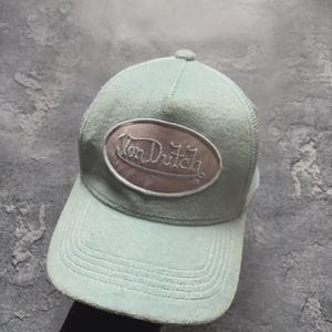 May include: A light blue Von Dutch baseball cap with a curved brim and mesh back. The cap features an oval patch with the brand name embroidered in a cursive font. The hat is a classic accessory.