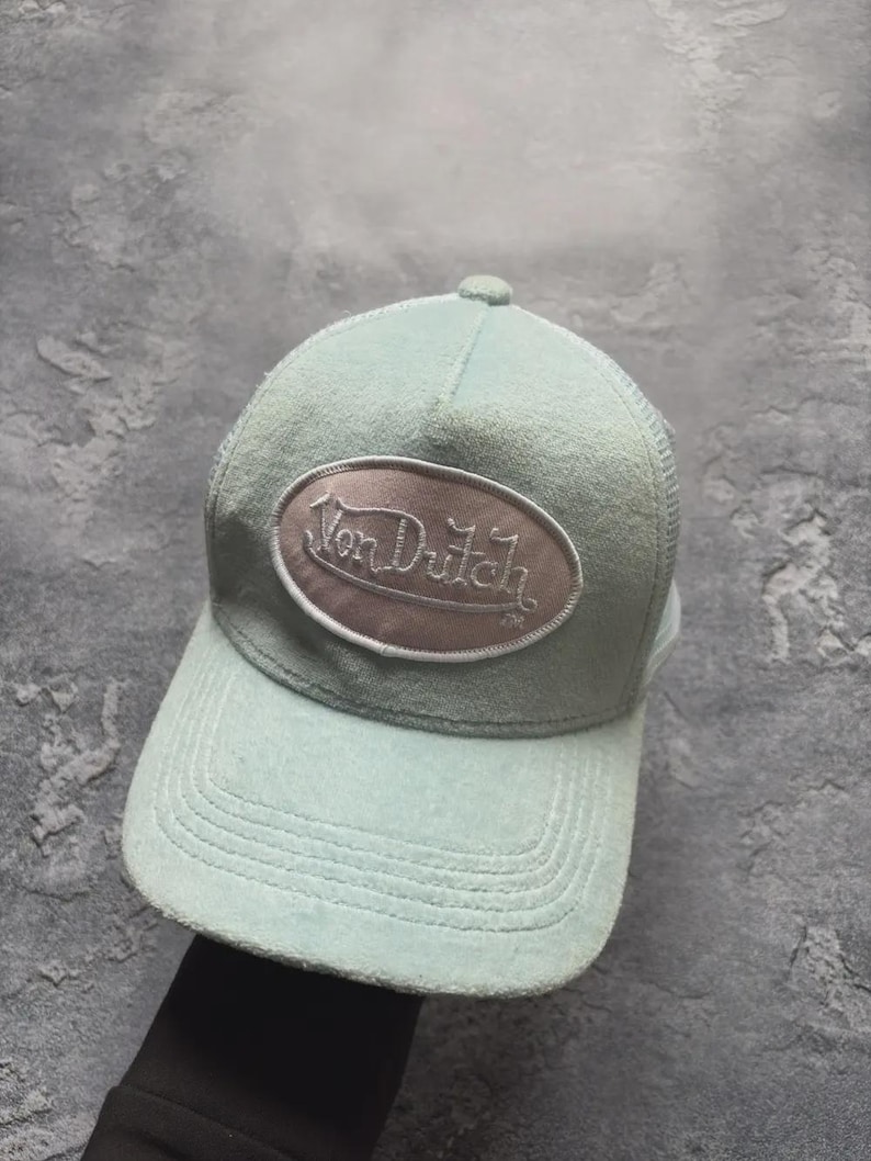 May include: A light blue Von Dutch baseball cap with a curved brim and mesh back. The cap features an oval patch with the brand name embroidered in a cursive font. The hat is a classic accessory.