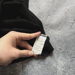 Może przedstawiać: Czarny podkoszulek z dzianiny wafelkowej. Tkanina ma teksturowany wz&oacute;r w kratkę. Widoczna jest metka z instrukcją pielęgnacji. Odzież wykonana jest z miękkiego, wygodnego materiału.