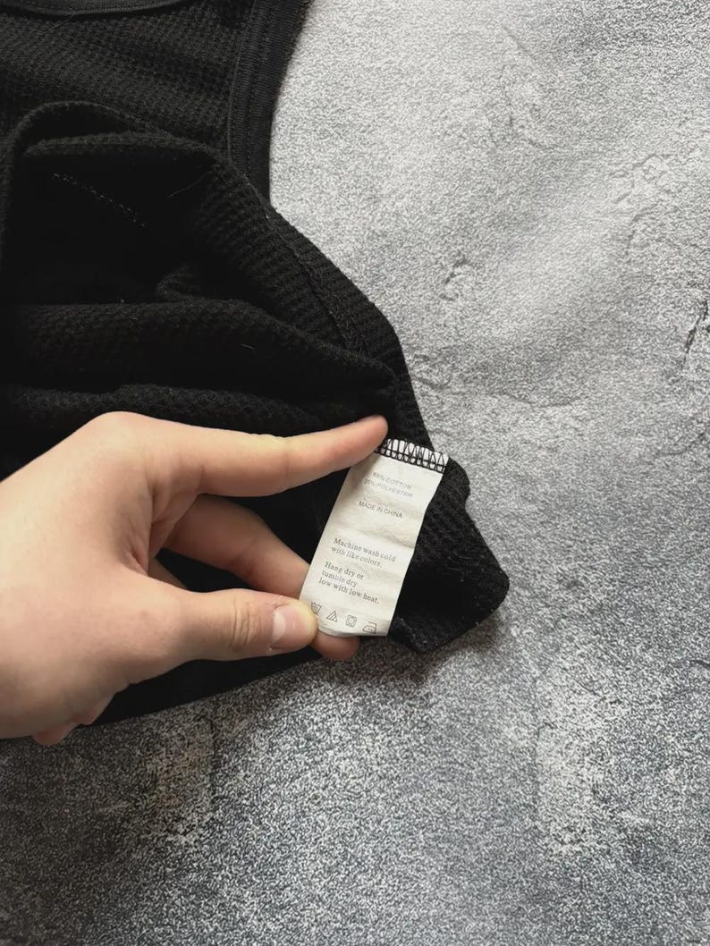 Może przedstawiać: Czarny podkoszulek z dzianiny wafelkowej. Tkanina ma teksturowany wz&oacute;r w kratkę. Widoczna jest metka z instrukcją pielęgnacji. Odzież wykonana jest z miękkiego, wygodnego materiału.