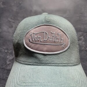 May include: A light blue cap with a curved brim and a matching top. The cap features an oval patch with the embroidered text "Von Dutch" in a script font. The hat is made of a soft, textured material.