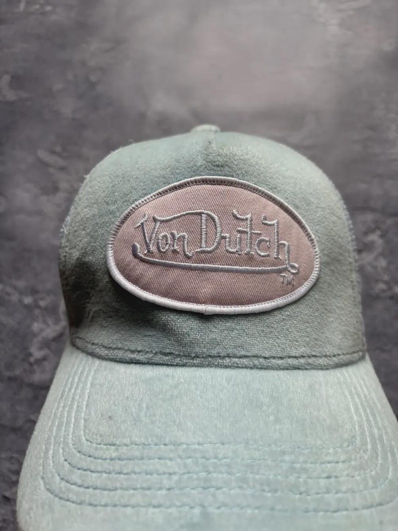 May include: A light blue cap with a curved brim and a matching top. The cap features an oval patch with the embroidered text "Von Dutch" in a script font. The hat is made of a soft, textured material.