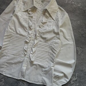 May include: White button-down shirt with long sleeves. The shirt features decorative ruffles down the front and intricate embroidered detailing on the shoulders and chest. The shirt has a classic collar and buttoned cuffs.