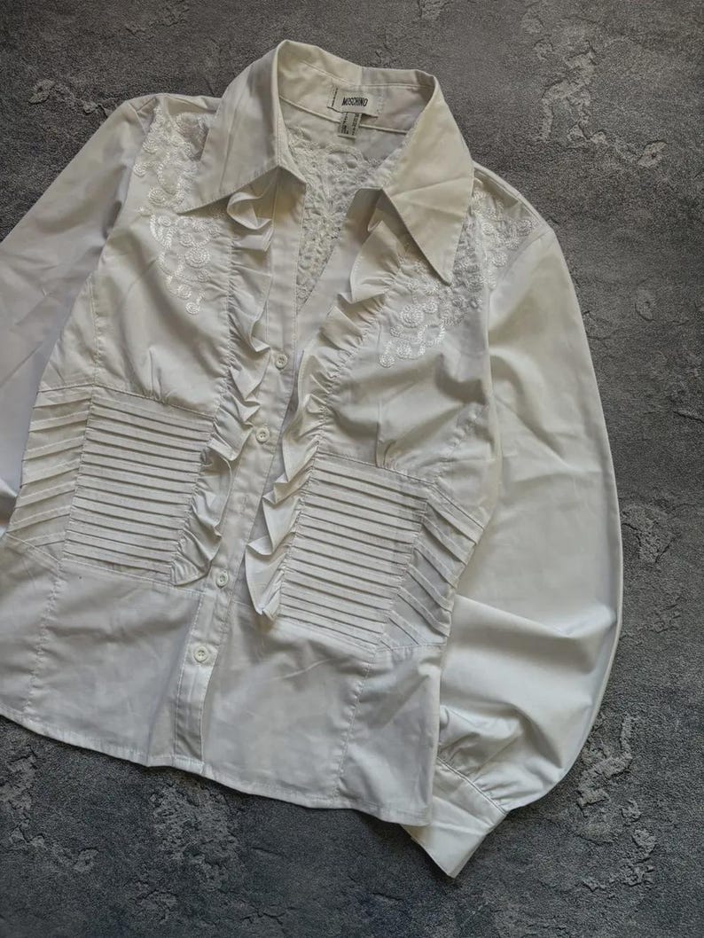 May include: White button-down shirt with long sleeves. The shirt features decorative ruffles down the front and intricate embroidered detailing on the shoulders and chest. The shirt has a classic collar and buttoned cuffs.