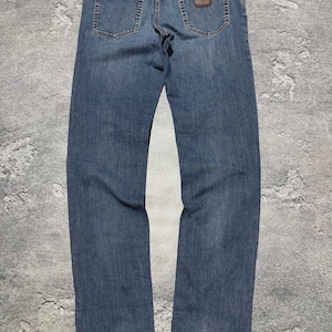 May include: Blue denim jeans with a classic five-pocket design. The jeans have a straight leg cut and a brown leather patch on the back waistband. The back pockets have a simple, rectangular shape.