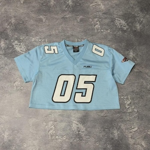 Può includere: Una maglia a maniche corte color azzurro con scollo a V. La parte anteriore presenta la scritta FUBU e il numero 05 in grandi cifre bianche con bordi neri. Cifre più piccole 5 e 0 appaiono sulle spalle e una toppa con logo è sulla manica.