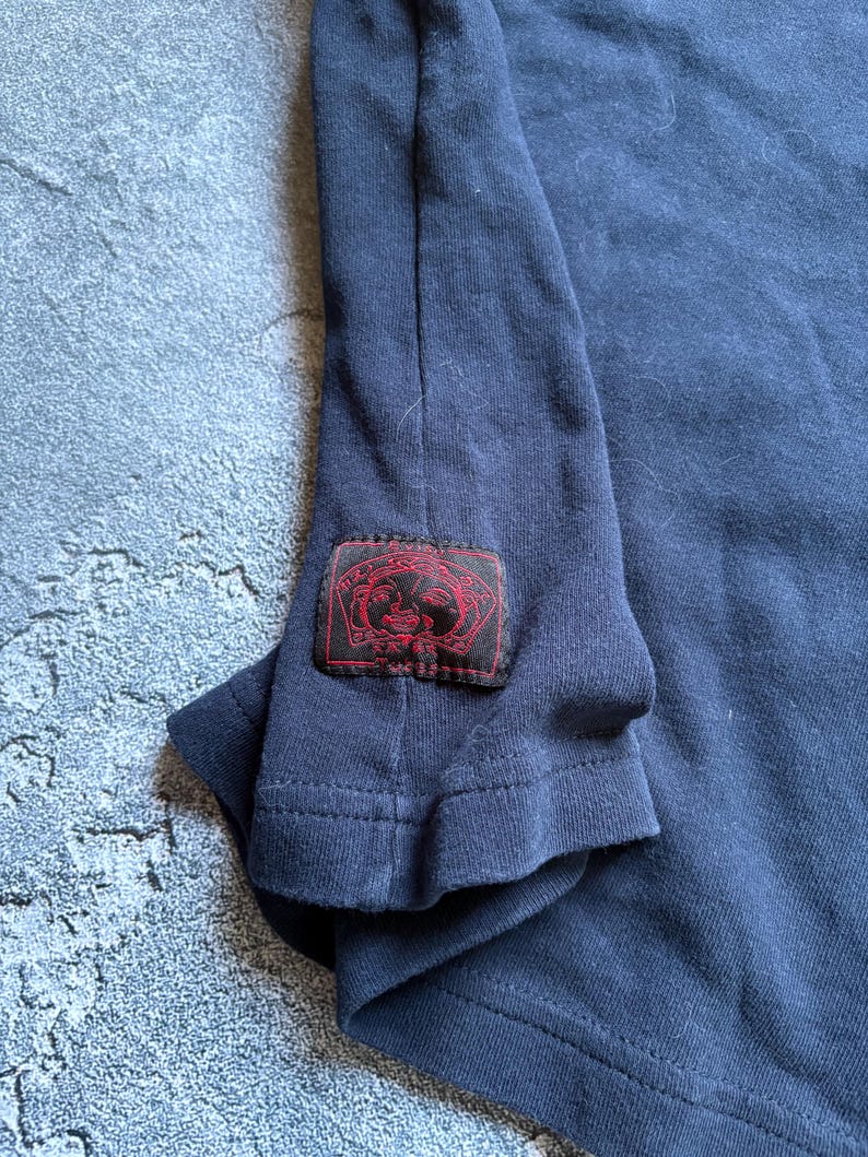 Puede incluir: Camiseta de manga larga azul marino con un parche rojo en la manga. El parche presenta un dise&ntilde;o con detalles rojos y negros. La camiseta est&aacute; hecha de un material suave y c&oacute;modo.